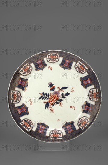 Platter with Imari decoration, Porzellanmanufaktur du Paquier Vienna; 1718–1744, imari style, Far Eastern flowers, Far Eastern style decorations. Round large platter with a multi-petaled collar edge with a wide cobalt trail with gilded diagonal latticework and a rhomboid 4-leaf rosette motif in the field. The cobalt trail features eight suspended cobalt valances with an inscribed bipartite tulip motif and symmetrical gilding, alternating with eight disc-shaped valances partially inscribed in a cobalt stripe, each with a peony blossom mortise and a suspended floral bifoliate palmette. The center of the mirror shows a loose composition of twigs and flowers, including peonies, surrounded by five loosely scattered brick-red flower twigs. On the reverse of the collar are two plum blossom twigs alternating with two cobalt grass reed? shoots, complemented by brick-red leaves with a flower. c. 1730, Vienna, Austria. Vessel; height 7.0 cm, width 42.0 cm., A round ceramic plate with a white cent...