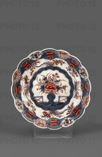Platter with Imari decoration, Kaiserliche Porzellanmanufaktur in Wien 1744–1864, imari style, Far Eastern style decorations. Round, bowl-shaped platter with a 16-petal edge and a 16-part lambrequin ornament trail symmetrically placed around the collar. On a wide cobalt-blue band around the edge are 8 brick-red peony flower motifs, each underpinned by a gold loop on the strap with a brick-red zigzag motif; alternating with the peony motifs are eight reserved disc-shaped fields at the edge with alternating brick-red plant arabesque motifs or cobalt–brick stylized floral-leaf motifs. In the lower part of the collar is a pattern of four flowered shoots with small flowers. A 16-petal mirror, bordered with a cobalt pattern with gilding, contains an inscribed motif of a bouquet in a cobalt vase flanked by two fences. At the edge on the reverse of the collar are 8 discs with a symmetrical brick-red floral and leafy motif, alternating with fields with a cobalt–brick-red stripe at the edge a br...