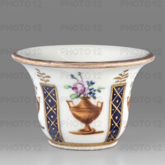 Toilet vessel for collecting tears or rubbing rouge?, Kaiserliche Porzellanmanufaktur in Wien 1744–1864. Bell-shaped porcelain vessel with a shallow circular recess; colorful decoration featuring four cobalt columns with gilded latticework alternating with four brown amphorae-vases containing floral arrangements; upper edge with gold borders. Identified as a vessel for collecting tears or for rubbing rouge?. 1807, Vienna, Austria. Toilet accessory. Height 3.5 cm; width 5.5 cm., A small white porcelain cup with a flared rim and a rounded base, decorated with painted floral motifs and ornamental panels; the rim and base feature a thin metallic-looking band in a warm gold color, and there are several vertical decorative panels in deep blue with gold lattice accents. Between the blue panels are painted brown urn shapes holding small bouquets with flowers in pink, purple, blue, and green leaves. The background is white and the overall palette includes white, gold, brown, deep blue, pink, pu...