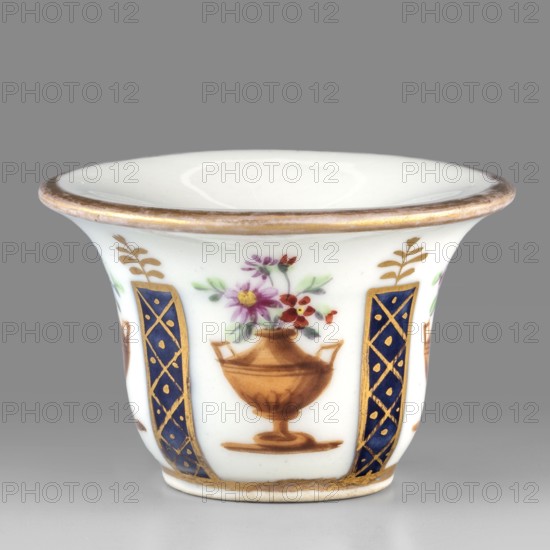 Toilet vessel for collecting tears or rubbing rouge?, Kaiserliche Porzellanmanufaktur in Wien 1744–1864. Bell-shaped porcelain vessel with a shallow circular recess; colorful decoration featuring four cobalt columns with gilded latticework alternating with four brown amphorae-vases containing floral arrangements; upper edge with gold borders. Identified as a vessel for collecting tears or rubbing rouge?. 1807, Vienna, Austria. Toilet accessory. Height 3.5 cm; width 5.5 cm. Maker: Kaiserliche Porzellanmanufaktur in Wien., A small white porcelain cup with a flared rim edged in gold shows painted decorations around its exterior: a stylized brown urn holding pink, purple, red, and yellow flowers with green leaves sits between vertical dark blue panels decorated with gold lattice and dot motifs, and small gold leafy sprigs appear above the panels; the cup rests on a short foot with a gold band near the base and the visible colors include white, gold, brown, dark blue, pink, purple, red, yel...