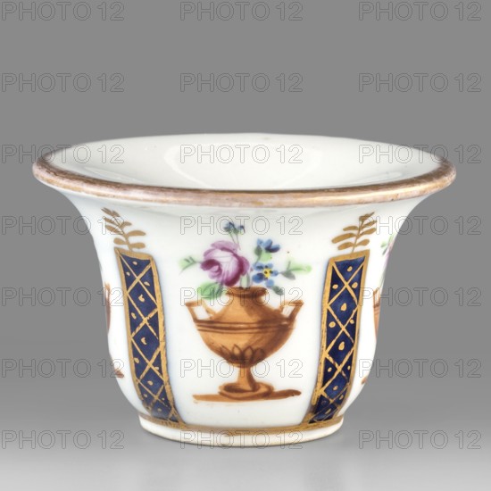Toilet vessel for collecting tears or rubbing rouge?, Kaiserliche Porzellanmanufaktur in Wien 1744–1864. Bell-shaped porcelain vessel with a shallow circular recess; colorful decoration with four cobalt columns with gilded latticework alternating with four brown amphorae-vases containing floral arrangements; upper edge with gold borders. Possibly identified as a vessel for collecting tears or for rubbing rouge. 1807, Vienna, Austria. Toilet accessory. Height 3.5 cm; width 5.5 cm. Maker: Kaiserliche Porzellanmanufaktur in Wien., A small white porcelain cup with a flared rim and a slightly footed base, decorated with painted motifs including a central brown urn holding a bouquet of flowers in shades of pink, blue, and green; the urn motif repeats around the cup and is separated by vertical panels in deep blue with gold lattice accents and additional gold leafy details; the rim and base edges show a gold band, and the background is plain white.