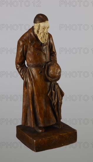 Figurine of a Jew, First Galician Factory of Artistic Faience and Terracotta Pacyków; 1912–1939. Figure of a Jew with sidelocks in a Crimean cap, dressed in a brown gabardine, holding a hat and umbrella in his left hand. Figure placed on a low plinth. 1st quarter of the 20th century, Pacyków. Terracotta/faience figurine; height 33.7 cm, width 13.0 cm, depth 8.0 cm., A standing figurine of an older man wearing a long coat and rounded cap, rendered in brown tones with a lighter beige face and beard; he holds a rounded hat and a draped cloth at his side, and the coat shows textured folds and buttons; the figure stands on a rectangular brown base against a plain light gray background.