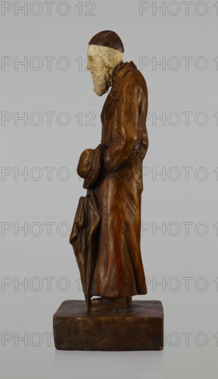 Figurine of a Jew, First Galician Factory of Artistic Faience and Terracotta Pacyków; 1912–1939. Figure of a Jew with sidelocks in a Crimean cap, dressed in a brown gabardine, holding a hat and umbrella in his left hand; figure placed on a low plinth. 1st quarter of the 20th century; Pacyków. Terracotta/faience figurine; height 33.7 cm, width 13.0 cm, depth 8.0 cm., A carved wooden figure of an elderly man shown in profile standing on a rectangular base, rendered in brown tones with lighter beige used for the face and beard; he wears a long brown coat and a darker brown cap, holds a brown hat in one hand and a folded umbrella or cloak in the other, and has a textured beard and hair with carved facial features against a plain light gray background.