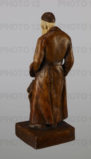 Figurine of a Jew, First Galician Factory of Artistic Faience and Terracotta Pacyków; 1912–1939. Figure of a Jew with sidelocks in a Crimean cap, dressed in a brown gabardine, holding a hat and umbrella in his left hand. Figure placed on a low plinth. 1st quarter of the 20th century. Pacyków. Figurine; height 33.7 cm, width 13.0 cm, depth 8.0 cm., A rear view of a carved figurine standing on a rectangular base, showing a long coat with a belt tied at the back and a cap on the head; the figurine has a textured beard and folded hands near the sides, and the surface shows a polished finish. Colors visible include brown on the coat, belt, cap, base, and parts of the figure; beige on the exposed head and beard; and gray in the background.