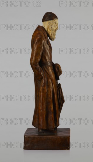 Figurine of a Jew, First Galician Factory of Artistic Faience and Terracotta Pacyków; 1912–1939. Figure of a Jew with sidelocks in a Crimean cap, dressed in a brown gabardine, holding a hat and umbrella in his left hand. Figure placed on a low plinth. 1st quarter of the 20th century, Pacyków. Terracotta/faience figurine; height 33.7 cm, width 13.0 cm, depth 8.0 cm., A carved wooden figure of an elderly man shown in profile standing on a rectangular base, wearing a long brown coat with visible folds and a belt, a dark brown cap, and holding a hat and an umbrella at his side; his exposed face and beard are a pale beige color with textured carving, and the overall colors visible are various shades of brown, dark brown, beige, and a neutral gray background.