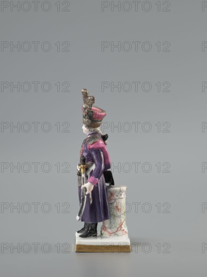Figurine of an officer of the Polish Guard, Koenigliche Porzellain Fabrique, Meissen 1710–1763, figurine; on a flat square base with gilded side edges, standing leaning against a low marbled column: a man in a purple kontusz with pink lapels, with epaulettes, a sabre strapped to the kontusz belt, a black map case slung on a belt and leaning against the column at the back; on his head is a high pink cap with black decorations and a high broken tail placed at the side. Defined signature on the bottom of the base as a Galician noble guardsman, in the Austrian service. 18th/19th century model/marking with the letter K 1-100 from ca. 1791. Saxony; Meissen., A small painted porcelain figure of a standing person wearing a tall hat with decorative elements, a long coat, and boots, shown in profile. The figure has visible colors including purple, pink, black, white, gold, and gray. The person holds an object resembling a sword with a metallic appearance and stands on a rectangular base with a g...