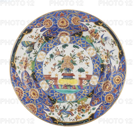 Plate, Karol Wolff faience factory; 1783–1800 maker, Sultan's service imitation, floral decorations, Far Eastern motifs, Stanislaw August Poniatowski King of Poland; 1732–1798, insects. Plate colorfully decorated with Far Eastern floral motifs; imitation of decoration from a Sultan's service produced by the royal factory in Belweder, late 18th century, Bielino, Poland; vessel; height 3.6 cm; width 25.0 cm, A round decorative plate with a central circular scene surrounded by patterned borders, featuring a floral arrangement in a vase at the center with a few stylized flowers and leaves in shades of yellow, pink, green, and blue, set on a base with brown and yellow tones; the inner ring around the central scene shows geometric and floral motifs in blue, gold, and white, and the outer rim is decorated with large floral designs and scrolling shapes in vivid blue, gold, brown, and touches of green and black, along with several small birds and butterflies in blue, green, and pink hues agains...