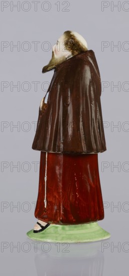 Three-part inkwell in the form of a monk figurine. Consisting of three movable parts: a movable head stopper, a body inkwell, and a lower part sandbox. Painted colorfully; the Capuchin habit is painted brown, the figure wears sandals on its bare feet, and holds a book in its hand. Engraved inscription Eidlbuss. Ca. 1800 18th/19th century. Prószków Slaski faience factory; 1763–1853. Province of Silesia, Silesia. Inkwell; material: faience/figurine. Height 18.5 cm, width 7.4 cm, depth 6.7 cm. Subjects: figurines, nuns, Capuchins order, inkwells., A glazed ceramic figurine of a standing person shown in profile, wearing a dark brown cloak over a red robe that extends to a pale green circular base; the head is pale with brown hair and a brown beard, and a pale face with subtle pinkish cheeks; a visible hand peeks from the cloak in a pale tone; the base sits on a reflective surface; the visible colors include brown, red, pale green, pale skin tones, pink, and black.