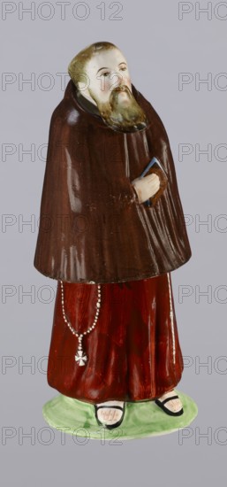 Three-part inkwell in the form of a monk figurine, ca. 1800 late 18th/early 19th century. Made at Prószków Slaski faience factory; 1763–1853. Consists of three movable parts: a removable head stopper, a body inkwell, and a lower part sandbox. Colorfully painted; Capuchin habit in brown, figure wears sandals on bare feet and holds a book. Engraved inscription Eidlbuss. Materials: faience. Function: inkwell. Dimensions: height 18.5 cm, width 7.4 cm, depth 6.7 cm. Region: Silesia Province of Silesia, 1742–1918., figurine, monk, nuns, Capuchins, figurines, inkwells., A ceramic figurine of a standing bearded man with a pale face and greenish-brown hair and beard wearing a dark brown cloak over a reddish-brown robe, holding a small object in one hand, with a string of beads hanging from the waist, standing on a light green base and wearing sandals showing pale feet; visible colors include brown, reddish-brown, green, pale skin tones, black, and white.