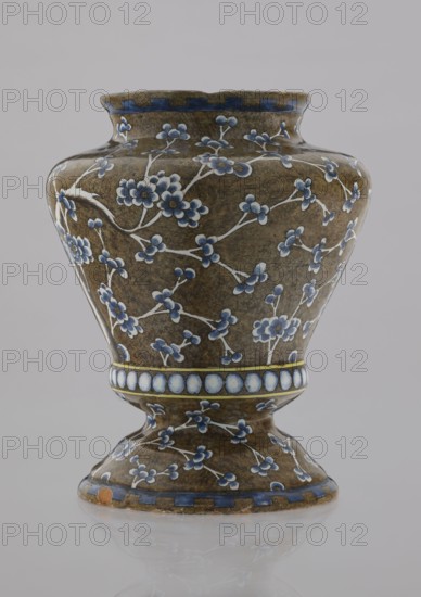 Vase by Karol Wolff fl. ca 1800; Bielino faience factory 1779–1800. A baluster-shaped Warsaw faience vase decorative vessel, profiled at the bottom with a short, slightly flared neck and a vaulted, profiled foot. Olive-brown body with a white, cobalt-shaded depiction of a plum tree whose bent, widely spread branch and network of flowery twigs cover the surface. Around the profiled belly is a row of white, shaded pearls; the rim bears a stylized teeth ornament. Chinoiserie/Far Eastern motifs, plant decorations. Inscribed/dated stylistically 1770–1800. Height 17.0 cm; width 13.2 cm. Origin: Warsaw, Poland., A ceramic vase with an hourglass-like silhouette sits against a pale gray background, its surface covered in a mottled brown glaze decorated with a pattern of white branches and small blue flowers and buds; the rim and base edges have a band of blue rectangles, and a central band around the narrowest part shows a row of pale blue circular medallions outlined in yellow, while the overa...