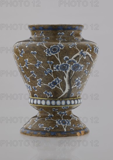 Vase, Karol Wolff fl. ca. 1800 and Bielino faience factory 1779–1800. A baluster-shaped faience vase with profiled base, short slightly flared neck, and vaulted profiled foot. Olive-brown body with a white, cobalt-shaded depiction of a plum tree whose bent, widely spread branch and network of flowery twigs cover the entire body. Around the profiled belly, a row of white, shaded pearls; at the rim, an ornament in the shape of stylized teeth. Chinoiserie/Far Eastern motifs, plant decorations. Warsaw, Poland, ca. 1770–1800. Decorative vessel; height 17.0 cm, width 13.2 cm., A ceramic vase with a rounded body and flared rim sitting on a pedestal base, decorated with floral motifs and branching stems in shades of blue and white against a brown background, with blue bands near the rim and base and a band of oval medallions encircling the waist; the surface shows glossy highlights and a few areas of worn or chipped brown glaze, and the vase is reflected faintly on a smooth pale surface.