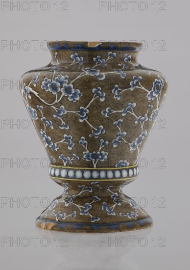 Vase, Karol Wolff fl. ca. 1800 author, Bielino faience factory 1779–1800 author; baluster-shaped vase with profiled bottom, short slightly flared neck, vaulted profiled foot; body olive-brown with white, cobalt-shaded depiction of a plum tree whose bent, widely spread branch covers the surface with a network of flowery twigs; row of white, shaded pearls around the profiled belly; edge ornament of stylized teeth; chinoiserie/Far Eastern motifs; plums; plant decorations; decorative vessel; 1770–1800; Warsaw, Poland; height 17.0 cm, width 13.2 cm, A ceramic vase with a rounded body and flared neck sitting on a pedestal base, covered in a brown mottled glaze and decorated with a pattern of small floral sprigs in shades of blue and white with pale stems, a band of circular motifs in white and blue encircling the midsection, and blue geometric border accents at the rim and base, with a small chip on the rim and another on the base, photographed against a plain light gray background.