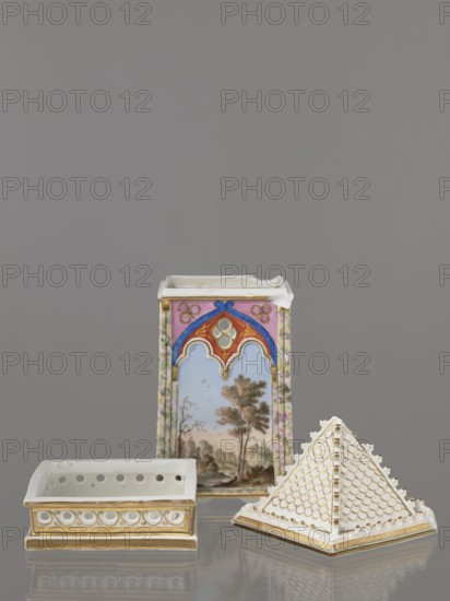 Night Lamp, Popow, Alexander porcelain manufacturer; 1806–1872 author, lamps, landscapes with architecture, neo-Gothic style, plant motifs. Lamp composed of three parts: base, lamp, and cover. Base with a horizontal row of holes on each side creating a decorative trail. Lamp in the form of a cube, in the neo-Gothic style; arcades in the shape of tracery painted with colorful landscapes: two with motifs of flowering bushes primrose and jasmine, floral motifs against the background of a landscape, two landscapes with an architectural motif. 19th century, Russia, Gorbunovo; lamp. Height 32.2 cm, width 13 cm., A still-life scene shows a group of decorative porcelain objects arranged against a plain gray background, including a tall rectangular vase with painted landscape panels and ornamental arches in colors of pink, blue, red, gold, green, and brown, a low rectangular dish with a white body and gold trim featuring circular relief decoration, and a pyramid-shaped cover with a white surfac...