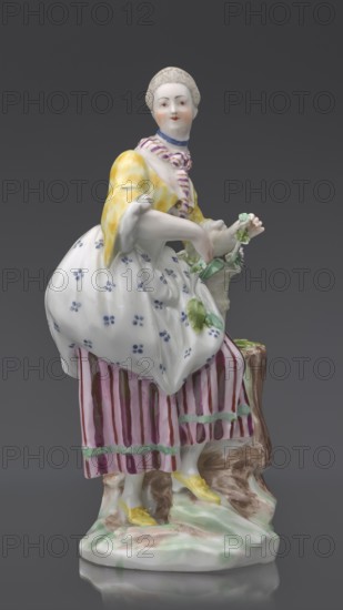 Figurine: gardener, Kaiserliche Porzellanmanufaktur in Wien 1744–1864 manufacturer, figurine, ca. 1770, Vienna, Austria; height 20 cm. On an irregular, rocky base, a young woman in a gardener's outfit leans her left arm on a tall basket of flowers standing on the base or a tree trunk?, holding twigs with leaves in her hands; leaning slightly to the left, with her left leg bent at the knee, resting on a rocky ledge. Dressed in a beetroot-striped skirt trimmed with green ribbon at the bottom, a tied-up apron with blue flowers, a short yellow jacket with flared sleeves, from under which the puffs of a white blouse peek out, a bodice with a gilded pattern, a cherry-striped scarf tied in a knot at the neck with long twisted ends falling to the waist and a blue ribbon with a bow at the neck; hair pinned up and combed smoothly., A porcelain figurine of a standing woman wearing a white skirt with small blue floral dots, a yellow jacket, and a striped skirt underneath in shades of pink and burg...