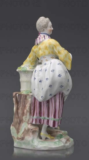 Figurine: gardener, Kaiserliche Porzellanmanufaktur in Wien 1744–1864 manufacturer, On an irregular, rocky base, a young woman in a gardener's outfit leans her left arm on a tall basket of flowers standing on a rocky base or a tree trunk?, holding twigs with leaves in her hands; leaning slightly to the left, with her left leg bent at the knee, resting on a rocky ledge. Dressed in a beetroot-striped skirt trimmed with green ribbon at the bottom, a tied-up apron with blue flowers, a short yellow jacket with flared sleeves, from under which the puffs of a white blouse peek out, a bodice with a gilded pattern, a cherry-striped scarf tied in a knot at the neck, with long twisted ends falling to the waist and a blue ribbon with a bow at the neck; hair pinned up and combed smoothly. ca. 1770, Vienna, Austria; porcelain figurine, height 20 cm, A porcelain figure of a woman standing beside a short tree stump, shown from the back and slightly in profile, wearing a yellow jacket with a marbled ap...