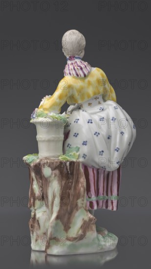 Figurine: gardener, Kaiserliche Porzellanmanufaktur in Wien 1744–1864; maker attribution Ryszard, Stanislaw Ryszard 1871–1955. On an irregular, rocky base, a young woman in a gardener's outfit leans her left arm on a tall basket of flowers standing on the base or a tree trunk?, holding twigs with leaves in her hands; leaning slightly to the left, left leg bent at the knee, resting on a rocky ledge. Dressed in a beetroot-striped skirt trimmed with green ribbon at the bottom, a tied-up apron with blue flowers, a short yellow jacket with flared sleeves revealing puffs of a white blouse, a bodice with a gilded pattern, a cherry-striped scarf knotted at the neck with long twisted ends falling to the waist and a blue ribbon bow at the neck; hair pinned up and combed smoothly. Ca. 1770, Vienna, Austria. Figurine, height 20 cm., A porcelain figurine viewed from behind shows a person with pale beige hair tied at the nape, wearing a yellow top with a mottled pattern and a striped neck scarf in p...