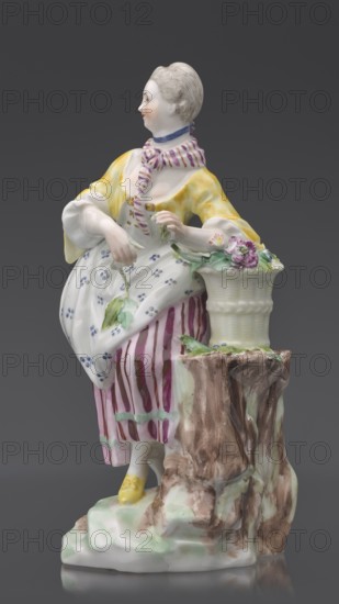Figurine: gardener, Kaiserliche Porzellanmanufaktur in Wien 1744–1864; on an irregular rocky base, a young woman in a gardener's outfit leans her left arm on a tall basket of flowers standing on the base or a tree trunk?, holding twigs with leaves in her hands; leaning slightly to the left, left leg bent at the knee, resting on a rocky ledge. Dressed in a beetroot-striped skirt trimmed with green ribbon at the bottom, a tied-up apron with blue flowers, a short yellow jacket with flared sleeves revealing puffs of a white blouse, a bodice with a gilded pattern, a cherry-striped scarf tied at the neck with long twisted ends falling to the waist and a blue ribbon with a bow at the neck; hair pinned up and combed smoothly. Ca. 1770, Vienna, Austria; porcelain figurine, height 20 cm., A painted porcelain figure of a seated woman wearing a yellow jacket, a white dress with small blue floral motifs, and a skirt with vertical pinkish-purple stripes, holding a green leaf in one hand and resting ...