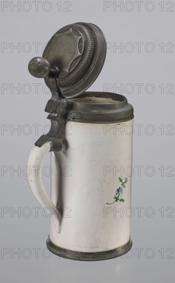 Cylindrical tankard in a pewter frame, Prószków Slaski faience factory; 1763–1853, maker/manufacturer inscriptions. Cylindrical tankard in a pewter frame. White faience, painted in colors. From the front: a barefoot woman sitting under a tree, blouse unbuttoned revealing her breasts; her head turned to the right, she supports with her right hand a small child, dressed in a short T-shirt, running towards her from the left — interpretable as an allegorical representation of Maternitas motherhood. The representation is painted with some freedom. Late 18th century, Province of Silesia 1742–1918, Silesia, Prószków Slaski. Vessel; height 24.0 cm, width 15.0 cm, depth 10.5 cm., A tall ceramic tankard with a hinged metal lid is shown against a neutral background; the tankard is primarily white with a glossy surface that shows some speckling and wear, a curved white handle on one side, and a small painted floral motif in blue and green on the body. The hinged lid and fittings are metal with a d...