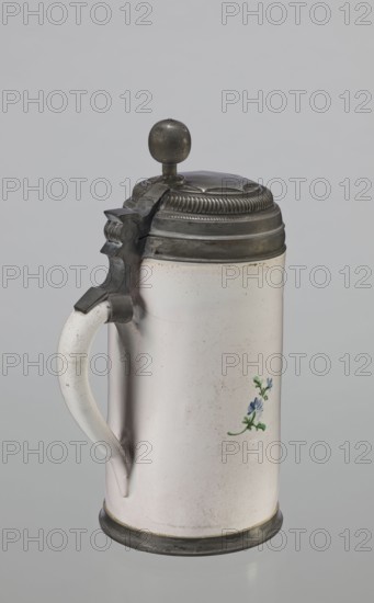 Cylindrical tankard in a pewter frame, Prószków Slaski faience factory 1763–1853. White, painted in colors. From the front, a depiction of a barefoot woman sitting under a tree, her blouse unbuttoned, revealing her breasts. The woman, head turned to the right, supports with her right hand a small child, dressed in a short T-shirt, running towards her from the left. It can be interpreted as an allegorical representation of Maternitas motherhood. The representation is painted with some freedom. Late 18th century, Silesia Province of Silesia, 1742–1918. Cylindrical tankard, vessel. Height 24.0 cm, width 15.0 cm, depth 10.5 cm., A cylindrical ceramic tankard with a pale off-white surface and a curved handle on the left is shown against a neutral gray background; it has a metal hinged lid topped with a round metal knob and decorative ridged banding, with matching metal fittings at the handle attachment and base, and a small painted floral motif in blue and green on the body.