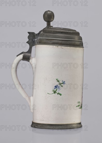 Cylindrical tankard in a pewter frame. White faience painted in colors. From the front, a barefoot woman sitting under a tree, her blouse unbuttoned, revealing her breasts; head turned to the right, she supports with her right hand a small child, dressed in a short T-shirt, running towards her from the left—interpreted as an allegorical representation of Maternitas motherhood. Painted with some freedom. Late 18th century, Prószków Slaski faience factory; 1763–1853, Province of Silesia 1742–1918, Silesia. Vessel. Height 24.0 cm; width 15.0 cm; depth 10.5 cm., A ceramic tankard with a hinged metal lid and thumb lever, shown in profile against a neutral background; the body is off-white with a glossy surface and small scattered imperfections, featuring a small floral painted motif in blue and green on its side and a hint of another motif at the edge, a curved off-white handle attached to the side, and metal bands at the base and around the top with a rounded metal finial on the lid; visib...
