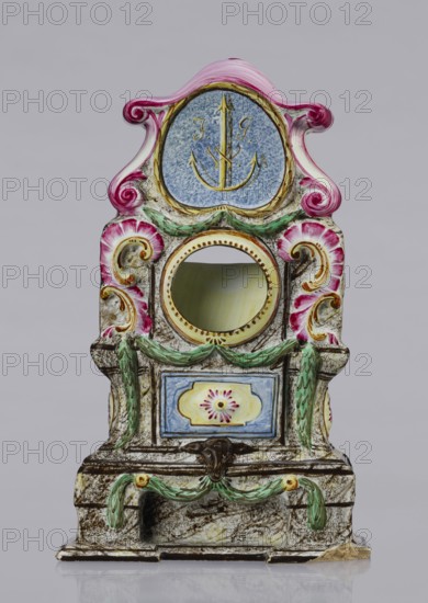 Clock setting, Prószków Slaski faience factory; 1763–1853. Clock setting in the shape of a late Baroque stove supported by a low base. Marbled, decoratively decorated, three-story, supported by a low base. In the central storey, above the pedestal with doors, is a circular recess for a watch framed by rocailles and laurel garlands. The volute-topped section features a shield with the monastic inscription JG/W against an anchor in the background. Painted colorfully in brown and purple tones, with yellow, green and blue accents. Late 18th century, Silesia Province of Silesia, 1742–1918, Prószków Slaski. Height 25.0 cm; width 15.1 cm; depth 5.0 cm., An ornate decorative object with a symmetrical, vertical form featuring curved and scroll-like shapes in pink and white at the top and sides, a central round opening with a yellowish inner rim, and a circular blue panel near the top with a gold-colored anchor-like motif; the piece shows painted garlands in green draped across the front, floral...