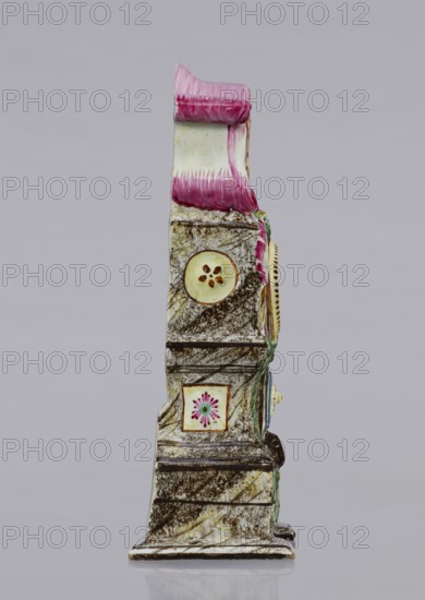 Clock setting, Prószków Slaski faience factory; 1763–1853. Clock setting in the shape of a late Baroque stove supported by a low base. Marbled, decoratively decorated, three-story, supported by a low base. In the central storey, above the pedestal with doors, is a circular recess for a watch framed by rocailles and laurel garlands. The volute-topped section features a shield with the monastic inscription JG/W against an anchor in the background. Painted colorfully in brown and purple tones, with yellow, green and blue accents. Late 18th century. Province of Silesia 1742–1918, Silesia, Prószków Slaski. Clock setting; height 25.0 cm, width 15.1 cm, depth 5.0 cm., A narrow vertical decorative object shown in profile with a glossy surface and a textured pattern of mottled brown and off-white, topped by a curved section painted in shades of pink and white, featuring a few circular and square inset motifs with floral-like designs in yellow, pink, green, and brown, and additional small accent...