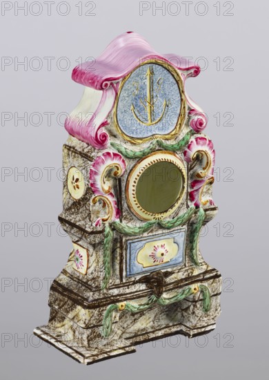 Clock setting, Prószków Slaski faience factory; 1763–1853. Clock setting in the shape of a late Baroque stove supported by a low base. Marbled, decoratively decorated, three-story, supported by a low base. In the central storey, above the pedestal with doors, is a circular recess for a watch framed by rocailles and laurel garlands. The volute-topped section features a shield with the monastic inscription JG/W against an anchor in the background. Painted colorfully in brown and purple tones, with yellow, green and blue accents. Late 18th century, Province of Silesia 1742–1918, Silesia, Prószków Slaski. Clock setting; height 25.0 cm, width 15.1 cm, depth 5.0 cm., An ornate decorative object with a sculpted form and a curved top painted pink and white, featuring a round central opening surrounded by gold and a blue circular panel above with a gold anchor-like motif, flanked by pink shell-like scrolls and green leafy garlands; the body has a marbled stone-like texture in beige and brown wi...