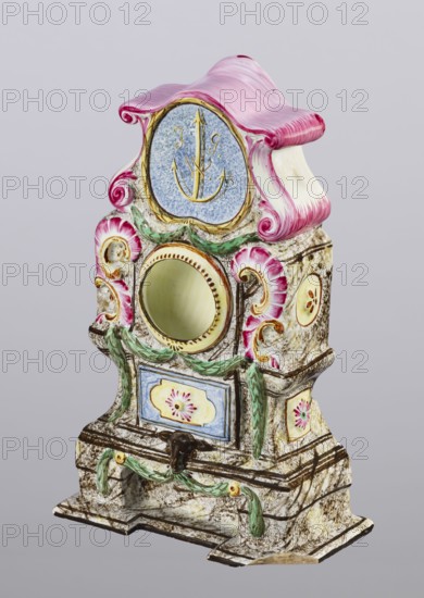 Clock setting, Prószków Slaski faience factory 1763–1853. Late Baroque stove–shaped clock setting on a low base; three-story, marbled and decoratively painted. Central storey above a pedestal with doors has a circular recess for a watch framed by rocailles and laurel garlands. Volute-topped section bears a shield with the monastic inscription JG/W against an anchor. Painted in brown and purple tones with yellow, green and blue accents. Late 18th century, Silesia Province of Silesia. Height 25.0 cm; width 15.1 cm; depth 5.0 cm., An ornate decorative object with a curving top in pink and white sits against a pale gray background; the piece features a round central opening with a pale yellow interior surrounded by painted gold accents, flanked by pink scroll-like motifs and green leafy garlands, and includes panels with floral medallion-like decorations in blue, yellow, and pink hues, all set on a mottled cream and brown base with additional green ribbon-like details and dark brown outlin...