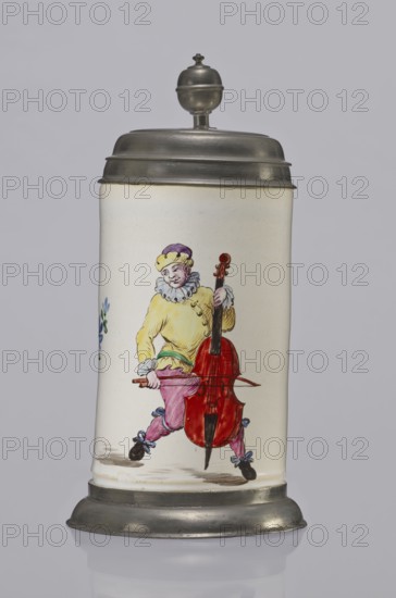 Tin-mounted tankard, Prószków Slaski faience factory 1763–1853. White cylindrical tankard in a pewter mount. On the front is a depiction of an obese musician playing a double bass, shown in a vigorous movement. The musician wears a yellow jacket, purple pantaloons, and a lilac hat with a yellow brim. Floral sprigs on the sides. On the tin cover engraved CGH/1811. Late 18th century. Province of Silesia 1742–1918. Dimensions: height 24.0 cm, width 15.0 cm, depth 9.5 cm., A tall cylindrical ceramic stein with a metal lid and base is shown against a plain light gray background; the ceramic body is off-white and features a painted figure of a person wearing a yellow top, purple hat, pink pants, blue bows on dark shoes, and holding a large red stringed instrument with a brown neck and black endpin, with a few small blue and green floral accents visible at the side.