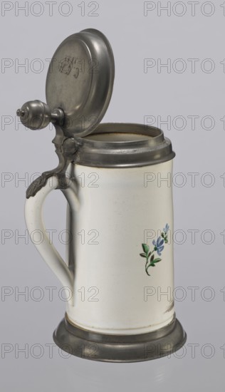 Tin-mounted tankard, Prószków Slaski faience factory 1763–1853. White cylindrical tankard in a pewter mount. On the front: an obese musician playing a double bass in vigorous movement, wearing a yellow jacket, purple pantaloons, and a lilac hat with a yellow brim. Floral sprigs on the sides. On the tin cover engraved CGH/1811. Late 18th century. Province of Silesia 1742–1918, Prószków Slaski. Vessel. Height 24.0 cm, width 15.0 cm, depth 9.5 cm., A tall ceramic stein with a hinged metal lid and base sits against a neutral background; the stein is primarily off-white with a smooth surface, a curved handle attached to the side, and a small painted floral decoration showing a few blue petals and green leaves; the metal lid, hinge, and base are a dull silver-gray with a slightly worn patina, and there are subtle darker gray shadows and highlights on the metal and white ceramic.