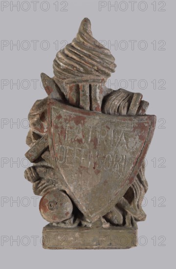 Fragment of a stove top, Jan III Sobieski King of Poland; 1629–1696, tiles, stoves. Triangular shield with the inscription PATRIAE DEFENSOR and panoplies. One of two panoplies flanking the model of the monument of Jan III Sobieski. 18th century, Poland. Tile; height 26.0 cm, width 16.5 cm., A weathered sculptural relief of a shielded emblem with a textured turban-like form at the top, flanked by draped cloth and curled ribbons, all carved in a worn surface showing layers of muted green, faded red, and grayish-beige paint; a round, disc-like object rests near the lower left of the shield, and the whole composition sits on a rectangular base, with visible chipping and flaking revealing underlying material.