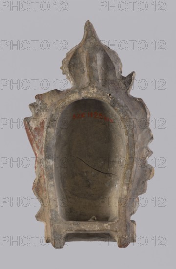 Fragment of a stove top by, depicting Jan III Sobieski King of Poland; 1629–1696. Earthenware tile showing a triangular shield inscribed PATRIAE DEFENSOR and panoplies; one of two panoplies flanking a model of the monument of Jan III Sobieski. 18th century, Poland. Tile, H 26.0 cm, W 16.5 cm., An upright hollow sculptural object with an arched recessed cavity, showing a weathered surface and rough edges, with decorative pointed and scalloped projections at the top and sides; the material appears stone or aged plaster with visible surface cracks and chips; colors visible include gray, beige, brown, and patches of muted red, with lighter and darker variations across the surfaces; the background is a plain light gray.