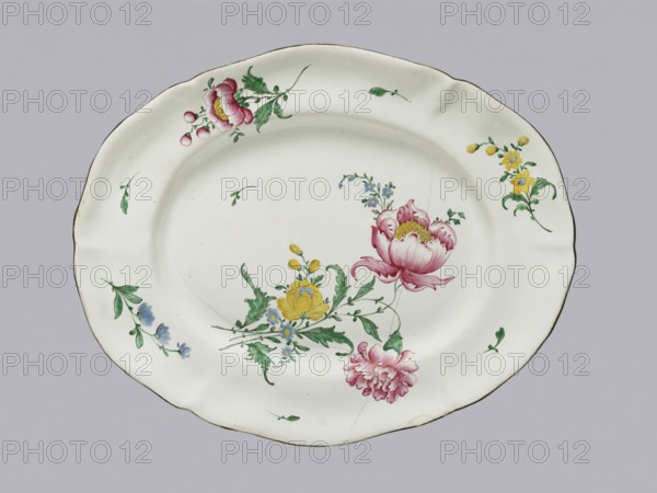Oval platter, Glinica faience factory 1771–ca.1870. Oval platter with a delicately modeled 6-petal collar. White, painted with colorful floral motifs. At the bottom is a bouquet with a large purple-pink peony and a carnation; below are yellow ranunculus and forget-me-nots. On the mirror are three small flower sprigs. On the collar are three larger floral arrangements of chicory, peonies with buds, and yellow flowers, interspersed with three small stems with leaves. Decoration executed with great care. Two large cracks and metal fastening clasps visible from previous conservation. 2nd half of the 19th century; 18th century; Province of Silesia 1742–1918, Silesia. Vessel; height 4.5 cm, width 33.0 cm, depth 41.0 cm., An oval white ceramic plate with a scalloped rim decorated with painted flowers, including a large pink peony-like bloom, a smaller pink carnation-like flower, yellow blossoms, and a few small blue flowers, accompanied by green leaves and stems; the rim has small scattered g...