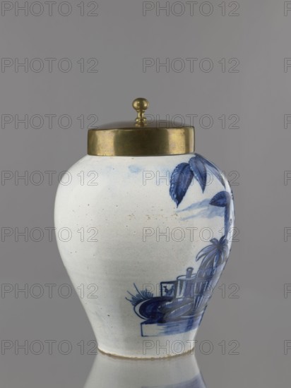 Tobacco jar with brass lid. Ovoid faience vase with a low, slightly parted neck, painted in cobalt on white. Genre scene depicting an Indian smoking a pipe under a tobacco bush and a vase labeled No. 17, arranged against a schematic seascape with two sailing ships. An Indian sits on a pedestal on which stands the labeled ovoid vase with lid; on the opposite side of the pedestal is a basket with a pineapple and a container. Similar vessels serving as tobacco jars were produced in the Netherlands throughout the 18th century. Inscriptions on the vase on the pedestal likely refer to the origin or type of tobacco. 18th century, Netherlands, Delft. Vessel: height 25 cm, width 21.5 cm; lid: height 7.6 cm, width 12.8 cm., A rounded ceramic jar with a domed metal lid topped by a small spherical finial sits against a plain gray background, the jar showing a white base color with blue painted decorative motifs including stylized leaves and a scene with architectural and plant forms, and the metal...