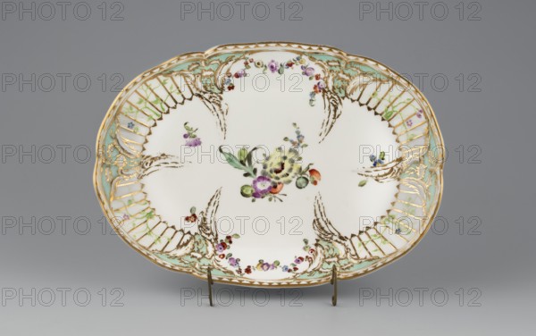 Vase base with Reliefzierat decoration; manufacturer: Königliche Porzellan-Manufaktur KPM, Berlin; ca. 1770–1780. Oval, bowl-shaped base with a relief of six triangular flame rocailles with celadon centers around the inner edge, separating four fields featuring a trellis of gilded stamens with an interwoven green branch with small flowers Reliefzierat. Two fields on the longer sides contain colorful floral garlands; the center mirror has a loose floral arrangement. Adjacent to the rocailles are scattered small flower sprigs. Around the edge of the base is a gilt strip separated by a half-cylinder. Maker association: Frederick II Frederick the Great, King of Prussia, 1712–1786. Origin: Berlin, Germany. Function: vessel., An oval ceramic platter sits on a small metal stand against a neutral gray background, primarily white with decorative painted floral motifs in the center and around the rim. The rim features gilded scrollwork and lattice-like cutout patterns accented with pale green an...
