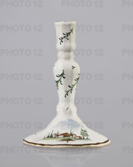 Candlestick, Prószków Slaski faience factory 1763–1853. Candlestick in the shape of a polygonal baluster supported on a wide, round conical base with a wavy edge. Painted in color. On the foot are two miniatures with a landscape with architecture; oblique projections of twigs on the shaft. Candlesticks; landscape miniatures; landscapes; revindication Henryków. Date: 1770–1783. Place: Prószków, Silesia, Province of Silesia. Dimensions: height 18.2 cm, width 13.0 cm., A white ceramic candlestick with a flared base and a slender, turned stem, decorated with a few green leafy sprigs on the stem and a painted pastoral scene on the base showing a group of small buildings, trees, and a low blue hill in the background; the rim of the base has a brown band. Colors visible include white, green, blue, brown, and touches of red.