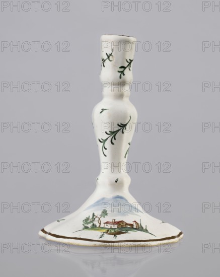 Candlestick, Prószków Slaski faience factory 1763–1853. Candlestick in the shape of a polygonal baluster supported on a wide, round conical base with a wavy edge. Painted colorfully; on the foot are two miniatures depicting landscapes with architecture, and oblique projections of twigs on the shaft. 1770–1783. Silesia Province of Silesia. Prószków Slaski. Porcelain/faience candlestick, height 18.2 cm, width 13.0 cm., A white ceramic candlestick with a rounded base and a slender, contoured stem decorated with small green leafy sprigs; the base shows a painted landscape scene with a small house, trees, and distant hills in muted greens, browns, blues, and a touch of red, and a thin brown rim around the edge.