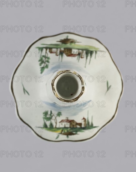 Candlestick, Prószków Slaski faience factory 1763–1853. Candlestick in the shape of a polygonal baluster supported on a wide, round conical base with a wavy edge. Painted in color. On the foot are two miniatures with landscapes featuring architecture; oblique projections of twigs on the shaft. Circa 1770–1783. Prószków Slaski, Silesia Province of Silesia. Height 18.2 cm; width 13.0 cm., A top-down view of a white ceramic cup and saucer set with a scalloped rim and hand-painted landscape scenes. The saucer shows a painted cottage scene with trees and a small building in green, brown, red, and blue hues, mirrored on opposite sides; there are a few green brushstrokes and a thin brownish-gold line around the scalloped edge. The cup sits centered on the saucer, showing its circular interior with a worn brownish edge and a muted pale green inside. The background is a flat light gray.