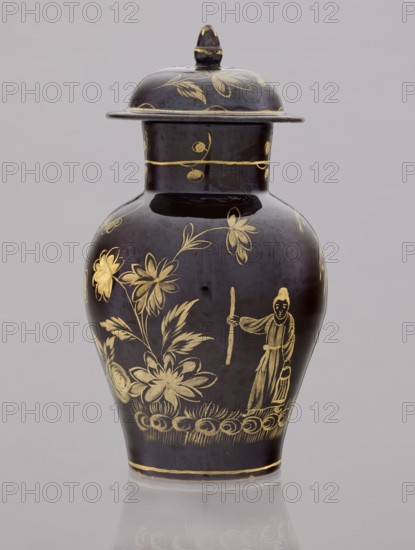 Kuan-type vase with cover, faience manufactory Bielino 1779–1800. Maker: Karol Wolff fl. ca. 1800. Chinoiserie—Chinese/Far Eastern motifs; Warsaw faience; unknown provenance. Low kuan-type vase in the shape of a bulging baluster with a cylindrical neck and a low domed cover with an acorn-shaped handle. Against a chocolate background a gold-painted scene: fragment of a garden with two chrysanthemum bushes, a peony bush, a palm tree, a rice fence, and between them the figure of a Chinese man with a bag and a walking stick. Around the neck is a thin simple plant twig; a painted flowered branch on the lid. Late 18th century, Poland. Vessel: height 19.5 cm, diameter 13.5 cm; lid: height 7.5 cm, diameter 10.5 cm., A dark ceramic lidded vase with a rounded body and a flared rim, decorated with gold-painted floral motifs and a gold-outlined figure holding a staff and a small object, the lid topped with a small finial, set against a pale gray background and reflected faintly on the surface belo...