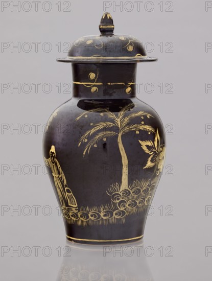 Low kuan-type vase with cover, faience Bielino manufactory, ca. 1779–1800, attributed to Karol Wolff fl. ca. 1800. Kuan-type vase in the shape of a bulging baluster with a cylindrical neck and a low domed cover with an acorn-shaped handle. Against a chocolate ground, gold-painted scene: fragment of a garden with two chrysanthemum bushes, a peony bush, a palm tree, a rice fence, and between them the figure of a Chinese man with a bag and a walking stick. Around the neck a thin plant twig; a painted flowered branch on the lid. Chinoiserie Chinese/Far Eastern motifs, late 18th century, Warsaw, Poland. Vessel: height 19.5 cm, diameter 13.5 cm; lid: height 7.5 cm, diameter 10.5 cm., A dark-colored lidded vase with glossy black and deep gray tones features gold decorative motifs including a stylized tree with slender leaves, a floral element, a standing human figure in flowing garments, and a band of looping ornamental patterns around the lower body; the lid and neck also show small gold cir...