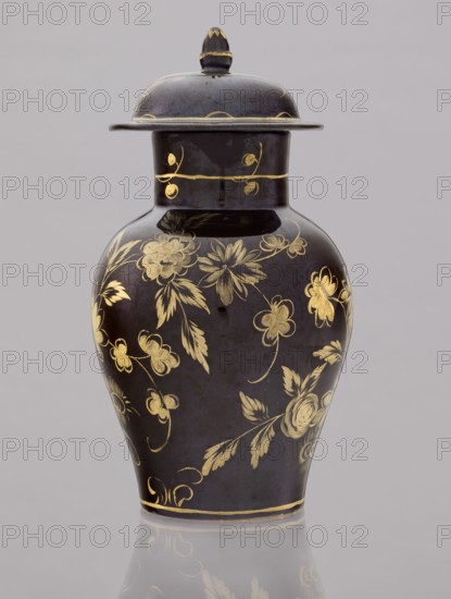 Kuan-type vase with cover, Bielino faience manufactory; 1779–1800. Maker: Wolff, Karol fl. ca 1800. Chinoiserie, Chinese/Far Eastern motifs; Warsaw faience; low kuan-type vase in the shape of a bulging baluster with a cylindrical neck and a low domed cover with an acorn-shaped handle. Against a chocolate background a gold-painted scene: fragment of a garden with two chrysanthemum bushes, a peony bush, a palm tree, a rice fence, and between them the figure of a Chinese man with a bag and a walking stick. Around the neck a thin simple plant twig; a painted flowered branch on the lid. Late 18th century. Place of origin: Warsaw, Poland. Vessel: height 19.5 cm, diameter 13.5 cm; lid: height 7.5 cm, diameter 10.5 cm., A dark vase with a lid sits against a light gray background, the vase and lid are a deep blackish color with shiny highlights, decorated with a floral pattern rendered in metallic gold; the gold designs include stylized blossoms, leaves, and curling vines distributed over the r...