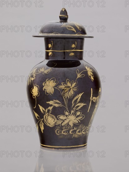 Kuan-type vase with cover, Bielino faience manufactory; 1779–1800. Maker: Karol Wolff fl. ca. 1800. Chinoiserie, Chinese/Far Eastern motifs, Warsaw faience, unknown provenance. Low kuan-type vase in the shape of a bulging baluster with a cylindrical neck and a low domed cover with an acorn-shaped handle. Against a chocolate background a gold-painted scene: a fragment of a garden with two chrysanthemum bushes, a peony bush, a palm tree, a rice fence, and between them the figure of a Chinese man with a bag and a walking stick. Around the neck is a thin simple plant twig; a painted flowered branch on the lid. Late 18th century, Poland. Vessel: height 19.5 cm, diameter 13.5 cm; lid: height 7.5 cm, diameter 10.5 cm., A lidded vase with a glossy dark surface decorated with floral and botanical designs in metallic gold tones; the vase has a rounded body that tapers toward the base and neck, a flared rim under the lid, and a domed lid with a small knob on top also decorated in gold; the visibl...