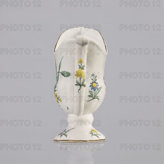 Nalewka, Prószków Slaski faience factory; 1763–1853, maker/manufacturer markings; floral decorations, locking, carnations, helmet-shaped nalewka with locking and fluted surface. Bowl base and vaulted foot modeled with four leaves. T-shaped handle. Colorful decoration: bouquet with a large purple carnation with three buds and three small yellow flowers; small projections of flowers and leaves. Brown borders along the handle, edge of the bowl, and foot. Dated 1770–1783. Region: Silesia Province of Silesia. Prószków Slaski. Vessel; height 19.0 cm, width 19.0 cm, depth 9.5 cm., A white ceramic pitcher with a handle and a short pedestal base is shown against a light gray background. The pitcher has hand-painted floral decorations in yellow, blue, green, and a touch of purple, with green stems and leaves. The rim and base edge have a thin gold trim. The pitcher’s surface shows subtle glaze variations and a glossy finish, and there is a soft reflection of the base on the surface beneath.