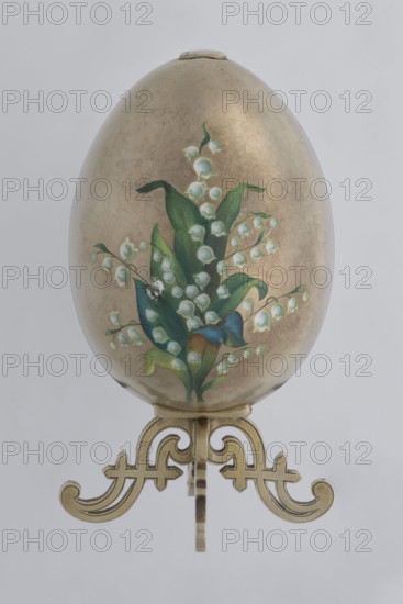 Easter egg, lilies of the valley and flowers motif; bequest of Jakub Ksawery Potocki noted 1863–1934. Silver egg, smooth, composed of two opening halves on a hinge, cut vertically; one part with an enameled bouquet of forget-me-nots; gilded interior with an octagonal basket for embedding a missing surprise. Egg supported on a four-legged stand. Date: 1890s, St. Petersburg. Decorative object. Height 11.4 cm; width 6.6 cm; depth 6.6 cm; weight 233.5 g., An oval object with a metallic gold surface sits on an ornate gold-toned stand; the front of the oval has a painted floral arrangement featuring green leaves and white bell-shaped flowers with hints of pale blue, and there are areas of darker green and a touch of blue and yellow in the lower leaves, all set against the warm gold background, with the stand showing intricate scrollwork in a matching gold color.