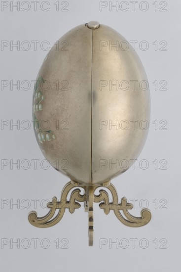 Easter egg, lilies of the valley, flowers, silver egg, smooth, composed of two opening halves on a hinge, cut vertically; one half with an enameled bouquet of forget-me-nots; gilded interior with an octagonal basket for embedding a surprise missing. Egg supported on a four-legged stand, 1890s, St. Petersburg, decorative object, height 11.4 cm, width 6.6 cm, depth 6.6 cm, weight 233.5 g, An egg-shaped metallic object resting on an ornate stand, shown from the front; the egg surface is smooth and reflective with a warm beige-gold tone and a slightly darker patina on one side, and a subtle seam running vertically down the center; the left side of the egg shows a small area of delicate painted floral accents in shades of green and pale white; the stand is decorative with scrolling curves and cutouts in a yellow-gold metal that matches the egg, set against a plain light gray background.