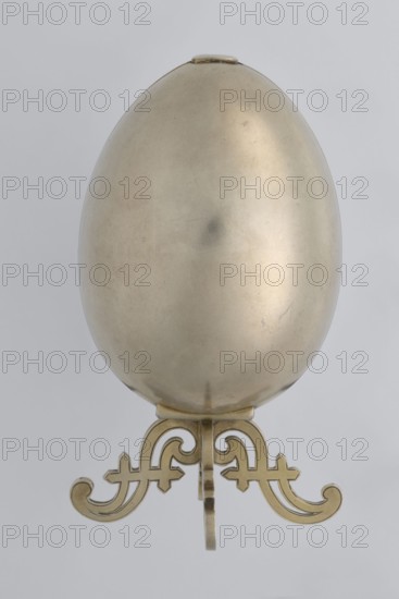 Easter egg, lilies of the valley, flowers, silver egg, smooth, composed of two opening halves on a hinge, cut vertically; one half with an enameled bouquet of forget-me-nots; gilded interior with an octagonal basket for embedding a missing surprise. Egg supported on a four-legged stand, 1890s, St. Petersburg, decorative object. Height 11.4 cm, width 6.6 cm, depth 6.6 cm, weight 233.5 g., A smooth metallic egg-shaped object with a pale gold finish placed upright on an ornate metallic stand; the egg surface shows subtle variations in sheen and a few faint blemishes, while the stand beneath is a decorative scrollwork form in a warmer gold tone against a light gray background.