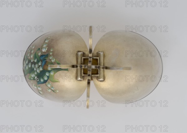 Easter egg, lilies of the valley, flowers; silver egg, smooth, composed of two opening halves on a hinge, cut vertically; on one part an enameled bouquet of forget-me-nots; gilded interior, with an octagonal basket for embedding a surprise missing. Egg supported on a four-legged stand, 1890s, St. Petersburg, decorative object. Height 11.4 cm, width 6.6 cm, depth 6.6 cm, weight 233.5 g., A smooth metallic object with a pale gold or brass tone sits against a light gray background; it consists of two rounded halves joined at a central square clasp with a cross-shaped metal hinge, and one rounded half shows a painted floral design in shades of green, blue, and white while the other half is plain and slightly mottled in the same pale gold color.