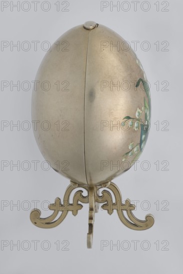 Easter egg, lilies of the valley and forget-me-nots decoration; silver egg, smooth, composed of two opening halves on a hinge, cut vertically; one part with an enameled bouquet of forget-me-nots; gilded interior with an octagonal basket for embedding a surprise missing. Egg supported on a four-legged stand. Decorative object, 1890s, St. Petersburg. Height 11.4 cm, width 6.6 cm, depth 6.6 cm, weight 233.5 g., An egg-shaped metallic object with a smooth, slightly worn surface resting on an ornate metallic stand; the egg shows a seam down its center and a small rounded hinge-like detail at its top, with a partial painted floral decoration showing green leaves and white blossoms on one side; the stand is decorative with scrolling cut-out shapes and a polished metallic finish in shades of gold and brass, and the background is a uniform light gray.