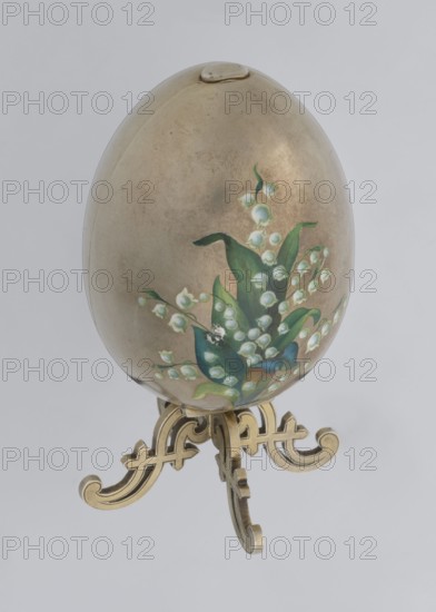 Easter egg, silver, smooth, composed of two opening halves on a hinge, cut vertically; on one part an enameled bouquet of forget-me-nots; gilded interior, with an octagonal basket for embedding a surprise missing. Egg supported on a four-legged stand. Decorated with lilies of the valley and flowers., Date: 1890s, St. Petersburg. Decorative object. Height 11.4 cm, width 6.6 cm, depth 6.6 cm, weight 233.5 g., An egg-shaped object with a metallic beige surface sits on an ornate stand; the egg features a painted floral cluster of bell-shaped blossoms and broad leaves in shades of green, white, and touches of blue, with the stand rendered in a gold color and the background a pale gray.