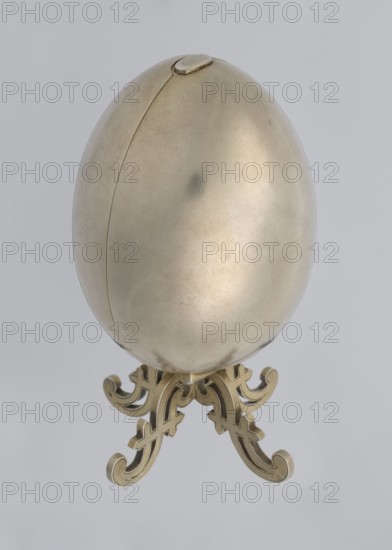 Easter egg, lilies of the valley and flowers motif, Easter egg, silver egg, smooth, composed of two opening halves on a hinge, cut vertically; on one part an enameled bouquet of forget-me-nots; gilded interior with an octagonal basket for embedding a surprise missing. Egg supported on a four-legged stand. 1890s, St. Petersburg. Decorative object. Height 11.4 cm, width 6.6 cm, depth 6.6 cm, weight 233.5 g., A smooth metallic egg-shaped object with a subtle seam and a small oval tab near its top sits on an ornate tripod stand; the egg surface shows gentle highlights, faint scratches, and a few darker spots, and the stand features a decorative scrollwork design. The visible colors are metallic gold and pale brass tones against a light gray background.